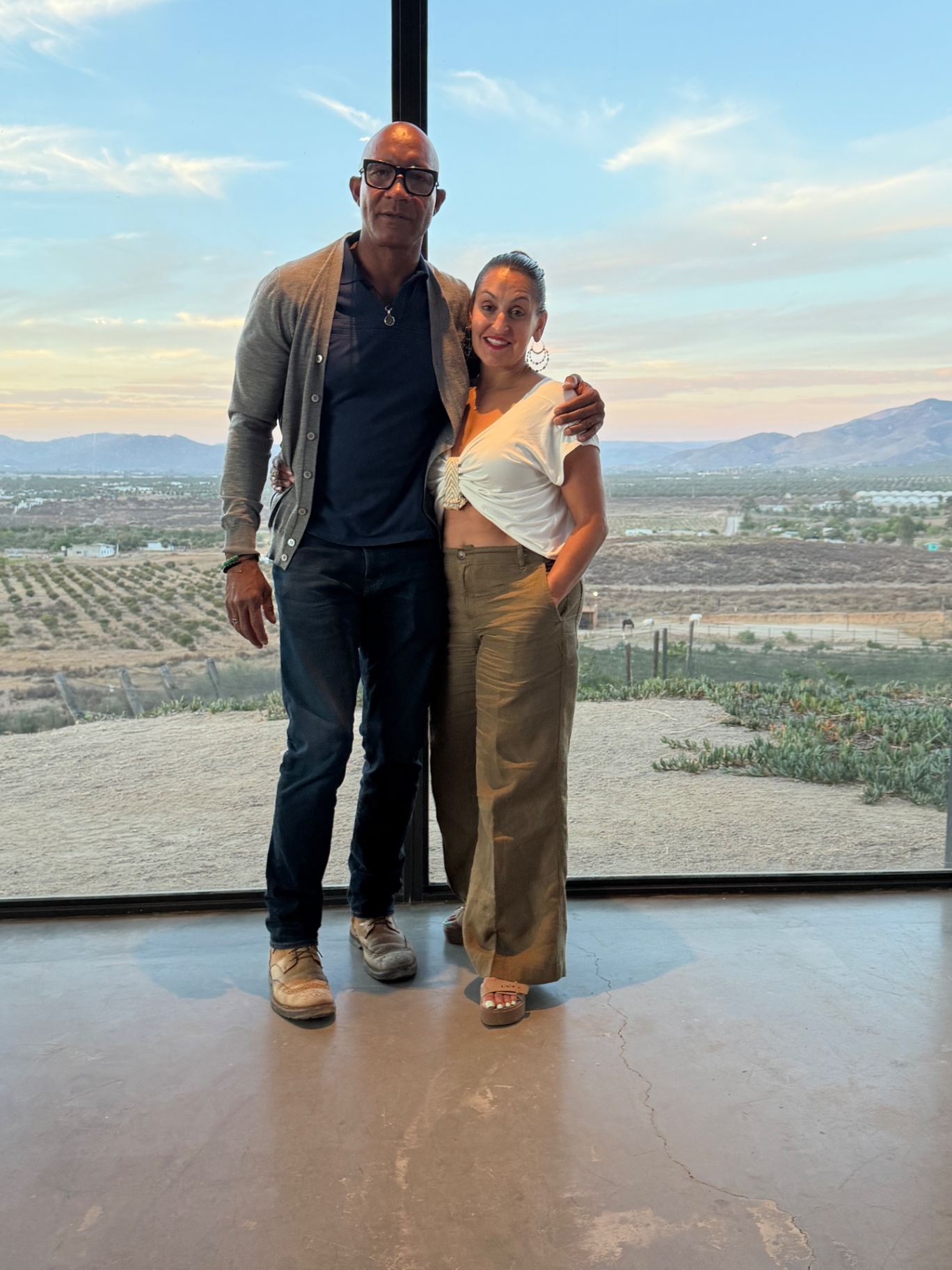 Sarah and Wilcil Jostein, founders of Antigua Valle, seated at a long table in Valle de Guadalupe, Baja California, with vineyards and hills at sunset.