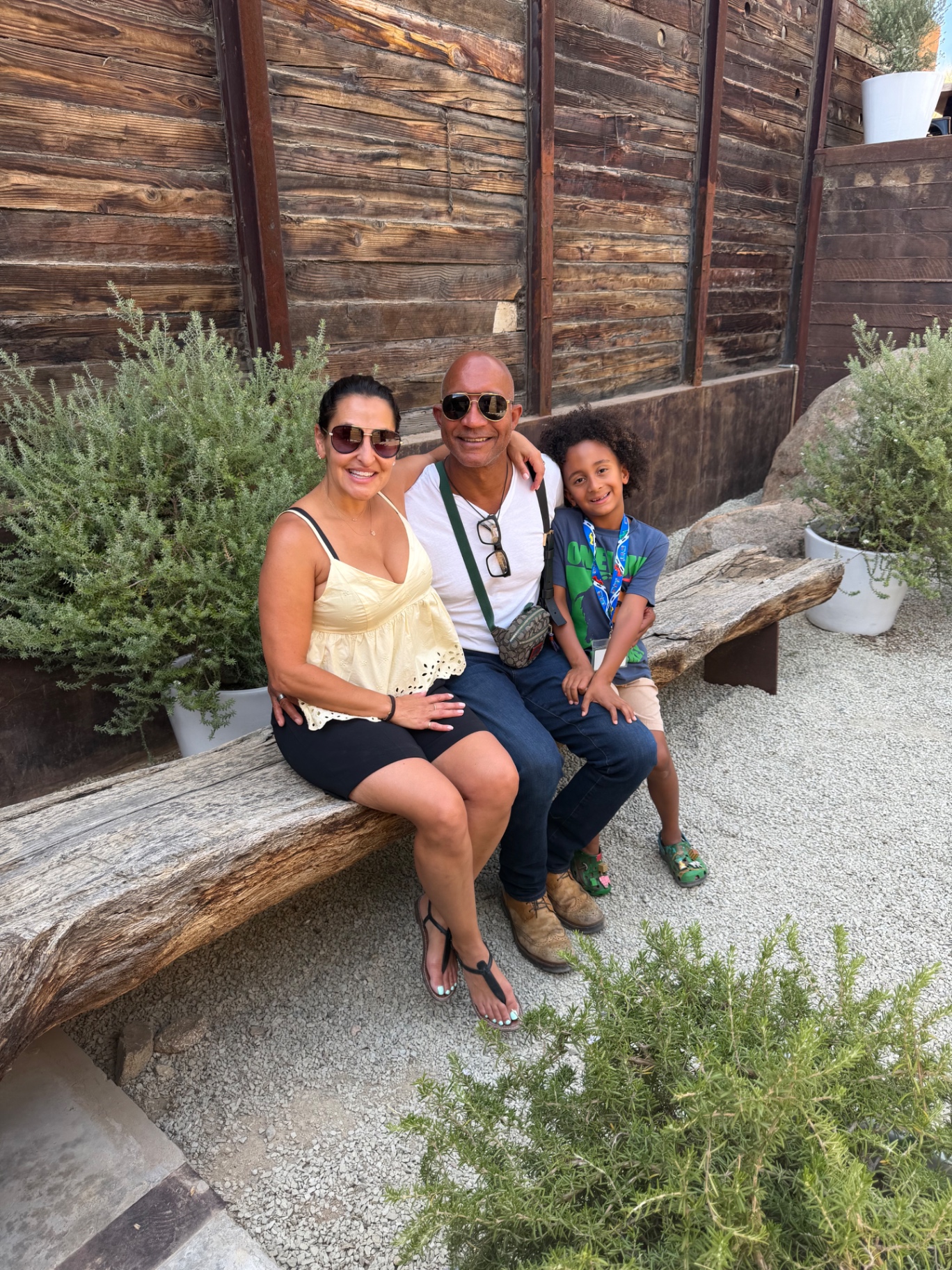 The Jostein family seated on a rustic wooden bench in Valle de Guadalupe — Sarah, Wilcil Joseph, and their son at a winery courtyard.